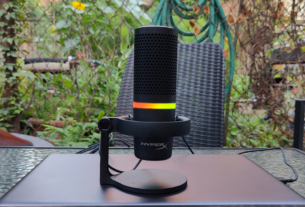 Microfone condensador USB HyperX DuoCast com anel de iluminação RGB colorido ativo, posicionado em um setup profissional de streaming e podcast.