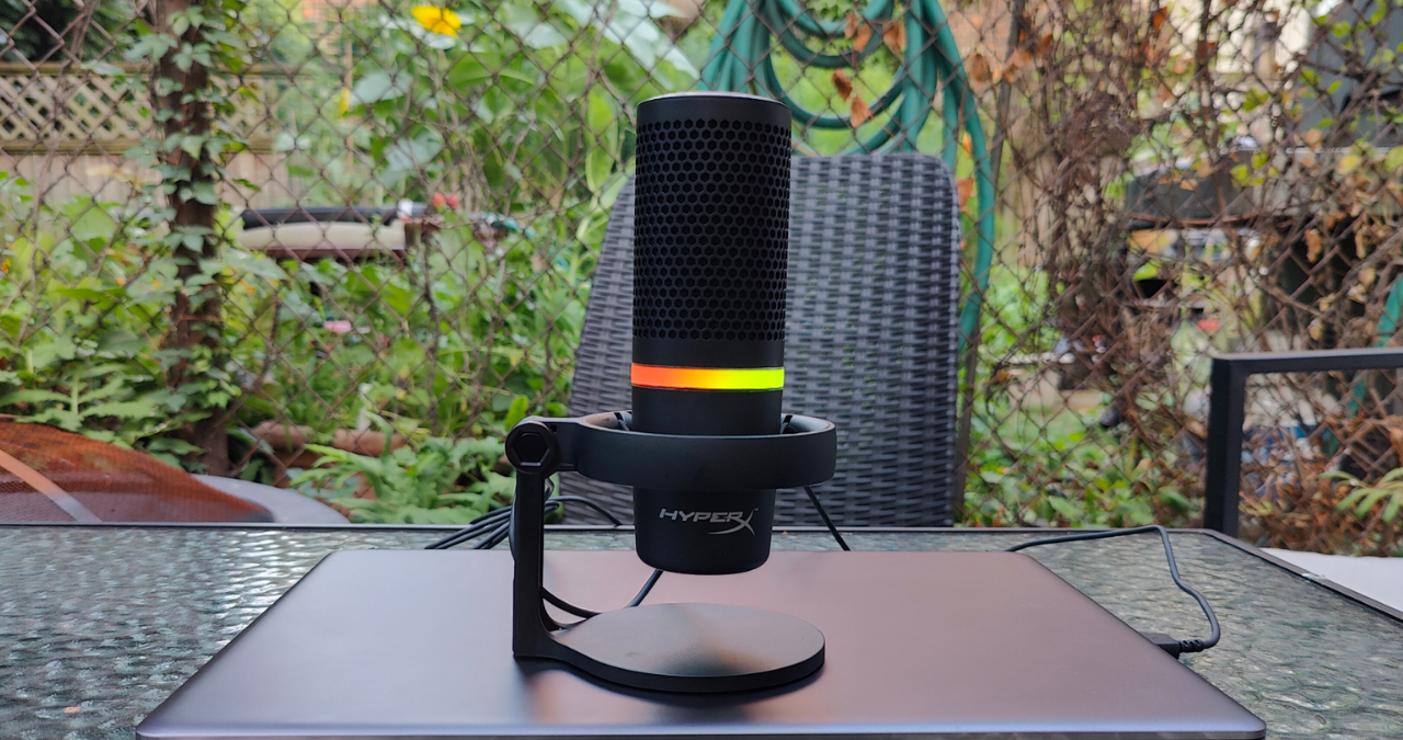 Microfone condensador USB HyperX DuoCast com anel de iluminação RGB colorido ativo, posicionado em um setup profissional de streaming e podcast.